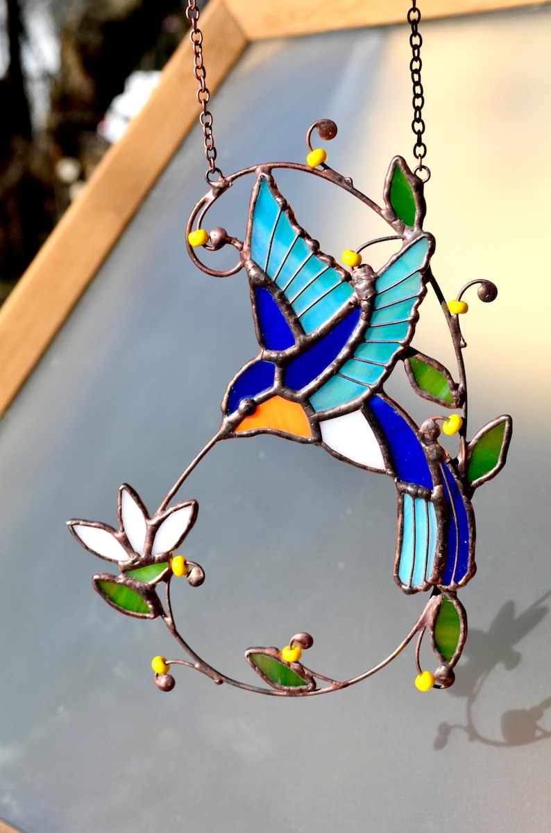 May include: A stained glass hummingbird with blue, green, and orange accents, perched on a copper wire branch with yellow beads. The hummingbird is in flight and has its beak extended as if it is feeding from a flower.