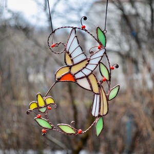 May include: A stained glass hummingbird with yellow, orange, and white wings, perched on a green and brown wire vine with red berries. The hummingbird is drinking nectar from a yellow flower.