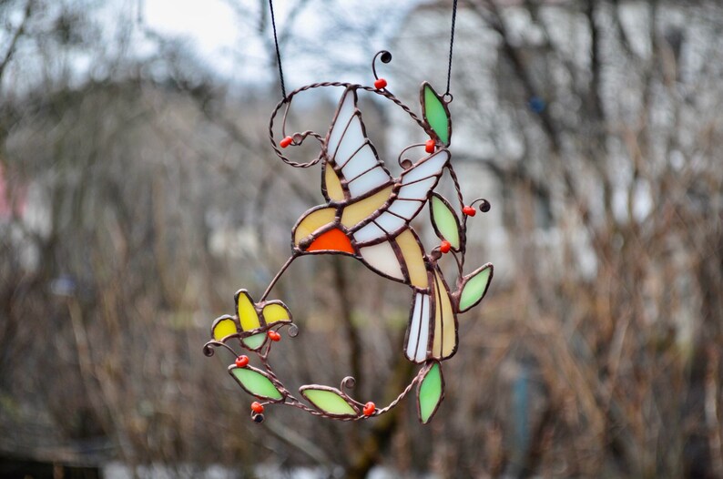 May include: A stained glass hummingbird with yellow, orange, and white wings, perched on a green and brown wire vine with red berries. The hummingbird is drinking nectar from a yellow flower.