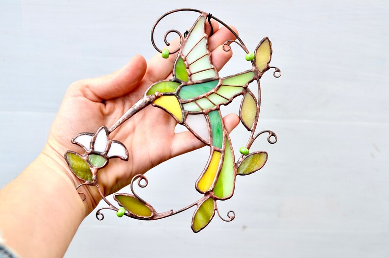 Pu&ograve; includere: Un colibr&igrave; in vetro colorato con pezzi di vetro verde, giallo e bianco. Il colibr&igrave; &egrave; appollaiato su una vite con foglie verdi e fiori bianchi.