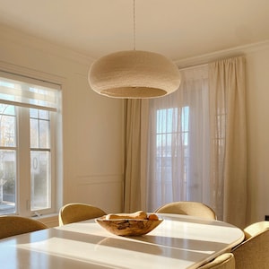 Op de afbeelding: Een witte gehaakte hanglamp hangt boven een witte ovale eettafel met zes stoelen. De stoelen zijn bekleed met een lichtbruine stof. Een houten kom met fruit staat op de tafel.