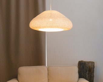 May include: A white woven pendant light hangs over a beige couch in a living room. The light is on and casts a warm glow on the couch and the surrounding area.