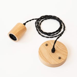 Puede incluir: Una lámpara colgante de madera con un cable trenzado negro. El accesorio incluye un portalámparas cilíndrico y una placa de techo redonda, ambos de madera clara. El cable negro está enrollado.