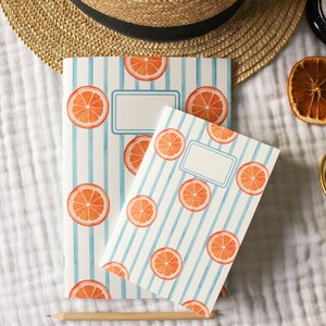 Puede incluir: Dos cuadernos con diseños de rodajas de naranja sobre un fondo a rayas blancas y azules. Los cuadernos tienen un área de etiqueta en blanco. También hay un sombrero de paja, una rodaja de naranja seca y pulseras doradas.