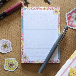 Può includere: Un blocco note floreale con un bordo blu e una penna grigia. Il blocco note ha uno sfondo bianco con carta rigata e un bordo floreale. Il testo "La carte du roman" è stampato nella parte inferiore del blocco note.