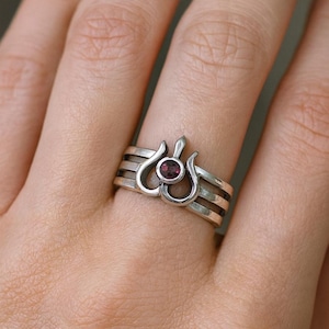 May include: A silver ring with a stylized floral design, featuring a small, round, dark red gemstone at the center. The ring has three bands and a decorative top element. The ring is on a person's finger.