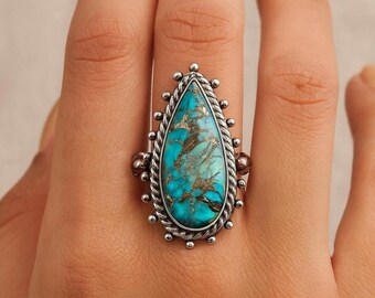 May include: A silver ring with a large, teardrop-shaped turquoise stone with brown veining. The stone is set in a silver frame with a twisted rope design and small silver beads. The ring is a statement piece.