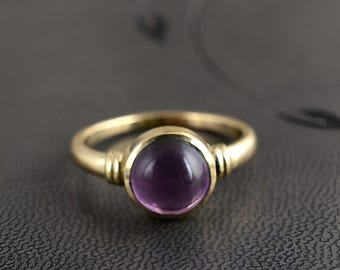 May include: A gold-colored ring featuring a large, round, purple gemstone. The ring has a simple band with decorative accents near the gemstone. The gemstone is a deep, rich purple color, and the ring appears to be made of metal.