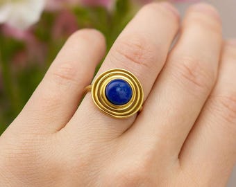 May include: A gold-colored ring featuring a deep blue, circular gemstone at its center. The ring's design includes concentric circles around the gemstone, creating a layered effect. The ring is shown on a person's finger, with a soft, blurred background.