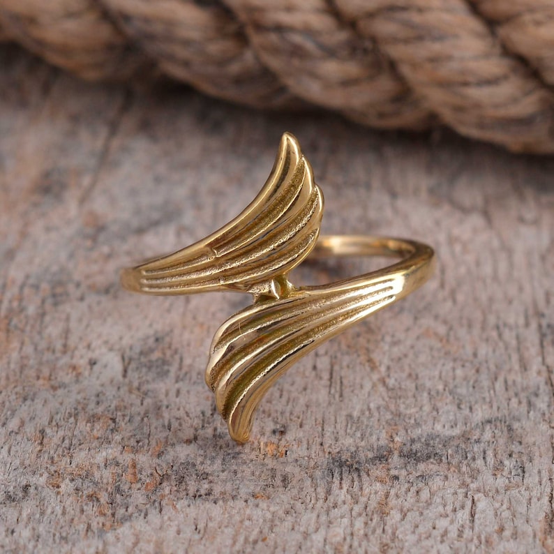 May include: A gold-colored ring with a wing-like design. The ring features two curved, layered wing shapes that meet in the center. The ring is set against a wooden surface.