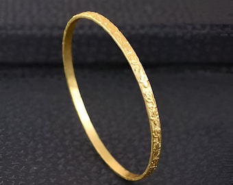 Puede incluir: Una pulsera dorada con un intrincado diseño en relieve. La pulsera es circular y parece estar hecha de metal. El patrón detallado cubre una parte de la superficie de la pulsera, agregando textura e interés visual. El fondo es de color oscuro y sólido.