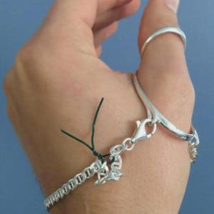May include: A silver bracelet with a chain-link design and a lobster clasp. The bracelet is connected to a ring on the thumb by a curved silver bar. A small charm hangs from the bracelet. The background is a solid blue.