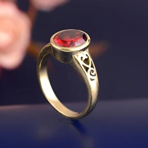 May include: A gold-colored ring with an oval, deep red gemstone. The ring has an ornate design on the band, with cut-out patterns. The gemstone is set in a bezel setting, and the ring is displayed against a dark blue background.