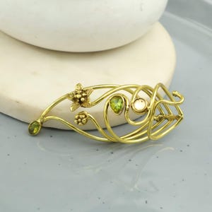 May include: A gold-toned hair barrette with an intricate, flowing design. It features small, pear-shaped green gemstones and a cluster of small, round white gemstones. The barrette also has floral and swirling details.