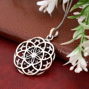 May include: A silver pendant necklace with an intricate, openwork floral pattern. The pendant is suspended from a fine chain. The necklace is shown on a brown surface with white flowers in the background.