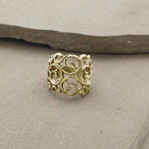 Könnte beinhalten: Ein goldfarbener Ring mit einem durchbrochenen Design mit wirbelnden, scrollartigen Mustern. Der Ring ist breit, mit einem Band, das etwa 2 cm breit ist. Der Ring ist vor einem neutralen Hintergrund platziert.