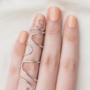 Könnte beinhalten: Ein silberner Ring mit abstraktem Design, der sich um einen Finger wickelt. Der Ring hat ein fließendes, geschwungenes Design mit einer Schlaufe oben und einer Reihe von Kurven. Die Hand ist mit pfirsichfarbenem Nagellack lackiert.
