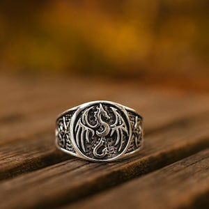 May include: A silver ring with a detailed dragon design. The ring features a circular emblem with a dragon in the centre, surrounded by intricate patterns. The ring is set against a blurred wooden background.
