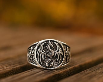 May include: A silver ring with a detailed dragon design. The ring features a circular emblem with a dragon in the center, surrounded by intricate patterns. The ring is set against a blurred wooden background.