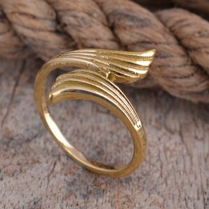 May include: A gold-colored ring with a wing-like design. The ring features a curved band that wraps around the finger, with the wing design extending outwards. The ring is made of a shiny, metallic material and is set against a wooden surface.