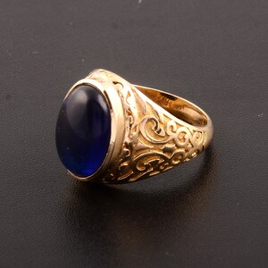 May include: A gold-colored ring with a large, oval, deep blue gemstone. The ring band features intricate, swirling, embossed patterns. The gemstone is smooth and polished, reflecting light. The ring is set against a dark background.