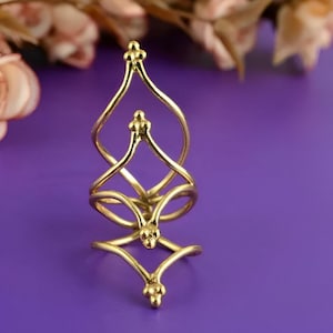 May include: A gold-colored, ornate ring with a unique, swirling design. The ring features multiple interconnected loops and decorative, rounded finials at the top and bottom. The ring is set against a purple background, with blurred flowers in the background.