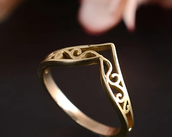 May include: A gold-colored ring with a V-shaped design. The ring features intricate openwork filigree patterns along the band and the V-shaped top. The ring is set against a dark background, highlighting its details.