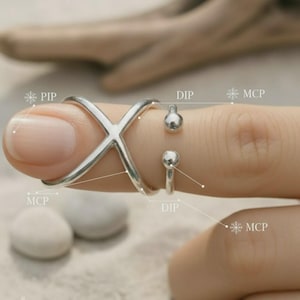 May include: A silver-colored ring with an X-shaped design and two small, round accents. The ring is worn on a finger, showcasing its intricate details and modern style. The ring's design is minimalist and elegant.