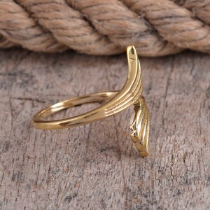 May include: A gold-colored ring with an open design. The ring features a curved, wing-like element with etched details. The ring is set against a wooden surface, with a rope in the background.
