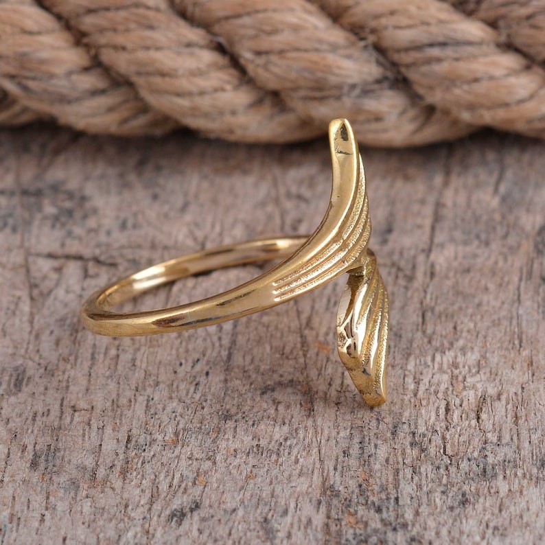May include: A gold-colored ring with an open design. The ring features a curved, wing-like element with etched details. The ring is set against a wooden surface, with a rope in the background.