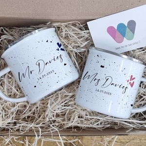 May include: Two white enamel mugs with black speckles and personalised text, "Mr Davies 14.07.2023" and "Mrs Davies 14.07.2023", in a white gift box with shredded paper. The box also contains a small card and colourful sweets.