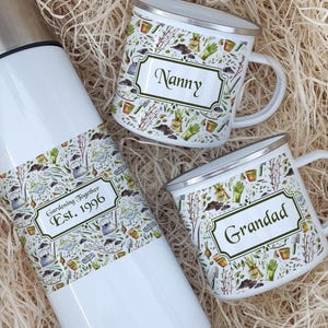 May include: A white thermos and two white mugs with silver rims, nestled in a bed of straw. The thermos has a decorative band with the text "Gardening Together Est. 1996". The mugs are labeled "Nanny" and "Grandad".
