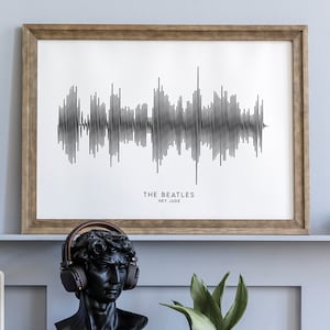 May include: A framed print of a black and white sound wave graphic with the text "THE BEATLES HEY JUDE" below. The print is on a white background and is framed in a brown wood frame. The print is hanging on a light blue wall. There is a black bust of a man wearing headphones, a white record player, a potted plant, and a white clock on a wooden table in front of the print.
