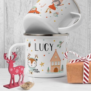 May include: Two white enamel mugs with silver rims, stacked. One mug features a cartoon of a girl, a car, and a penguin. The other mug has the name "LUCY", a deer, and a house. A red reindeer decoration and a wrapped gift are also present.