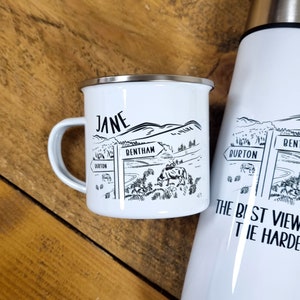 K&ouml;nnte beinhalten: Eine wei&szlig;e Emailletasse mit einer Schwarzwei&szlig;illustration einer l&auml;ndlichen Szene mit einem Wegweiser, auf dem "Bentham" steht. Die Tasse hat einen schwarzen Henkel und den Namen "Jane" auf der Seite.