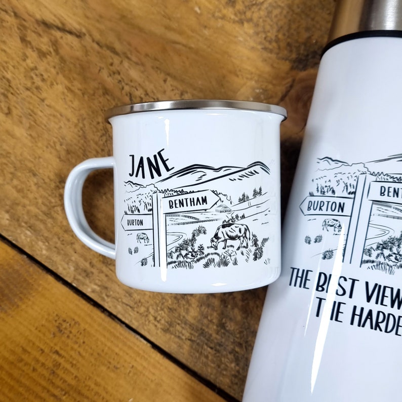 K&ouml;nnte beinhalten: Eine wei&szlig;e Emailletasse mit einer Schwarzwei&szlig;illustration einer l&auml;ndlichen Szene mit einem Wegweiser, auf dem "Bentham" steht. Die Tasse hat einen schwarzen Henkel und den Namen "Jane" auf der Seite.