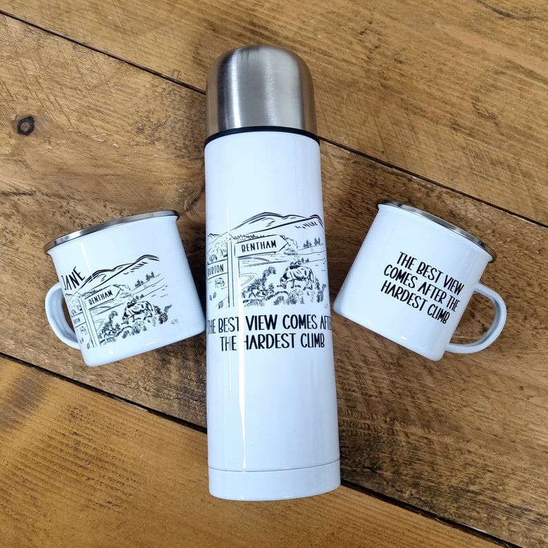 K&ouml;nnte beinhalten: Zwei wei&szlig;e Emailletassen mit schwarzem Text und Illustrationen einer Bergkette und dem Text "The best view comes after the hardest climb". Eine wei&szlig;e Thermoskanne mit silbernem Deckel und dem gleichen Text und der gleichen Illustration befindet sich in der Mitte.