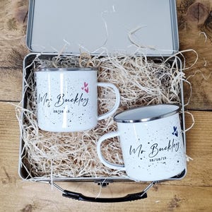 May include: Two white enamel mugs with black speckles and silver rims, each inscribed with "Mr. Buckley" and a date. The mugs are inside a metal tin filled with straw, ideal for gifting. Pink and blue heart accents are also present.