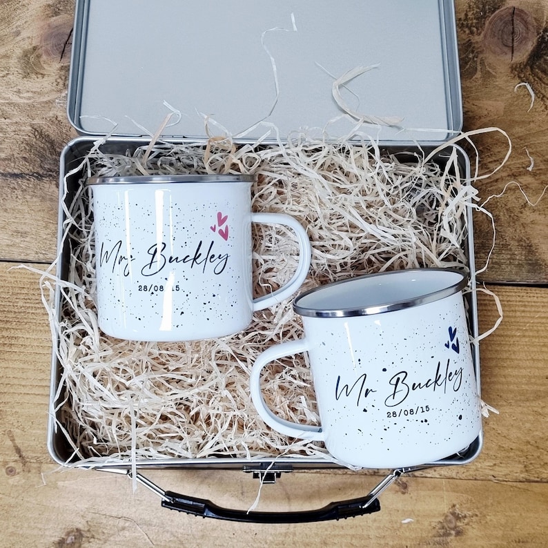 May include: Two white enamel mugs with black speckles and silver rims, each inscribed with "Mr. Buckley" and a date. The mugs are inside a metal tin filled with straw, ideal for gifting. Pink and blue heart accents are also present.