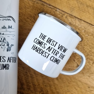 K&ouml;nnte beinhalten: Wei&szlig;e Emaille-Campingtasse mit schwarzem Rand und Henkel. Die Tasse tr&auml;gt eine schwarze Aufschrift mit dem Text "Die beste Aussicht kommt nach dem schwierigsten Aufstieg."
