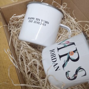 Pode incluir: Duas canecas de esmalte branco em uma caixa cheia de papel picado. Uma caneca diz "Happy Tin Years Est 23.02.2013" e a outra caneca diz "Mrs Jordan" com desenhos florais.