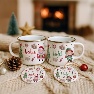 May include: Two white enamel mugs with gold rims, each featuring a Christmas design and a name. One mug has a Santa Claus illustration, the other an elf. Matching ornaments are also present.