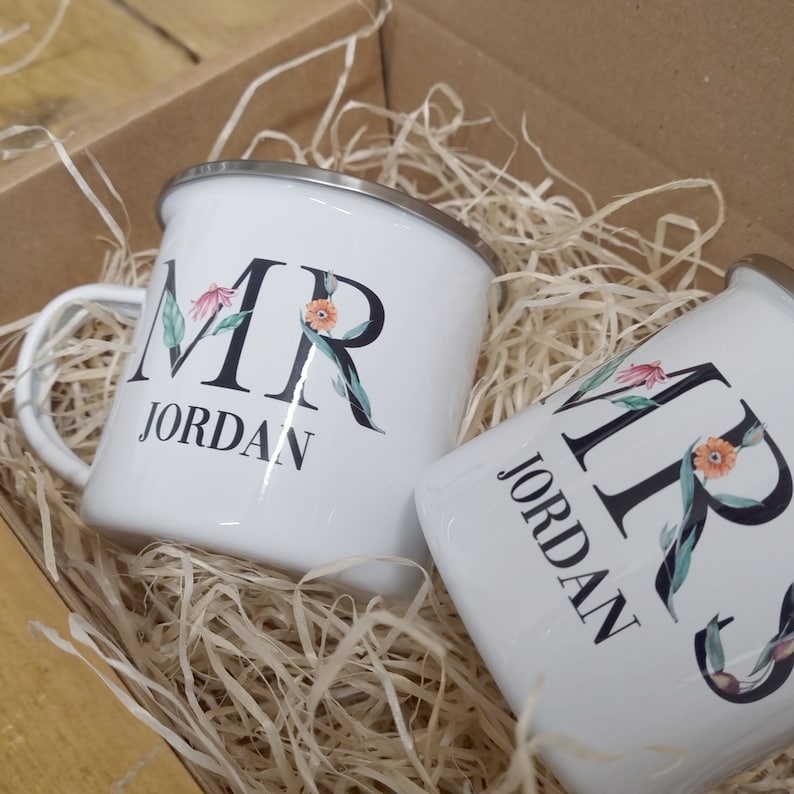 Pode incluir: Duas canecas de esmalte branco com letras pretas e detalhes florais. As canecas dizem "MR JORDAN" e "MRS JORDAN".