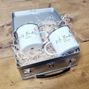 May include: Two white enamel mugs with black speckles and the names "Mrs Buckley" and "Mr Buckley" in script, along with the date "28/08/15". The mugs are inside a silver metal lunchbox filled with straw.