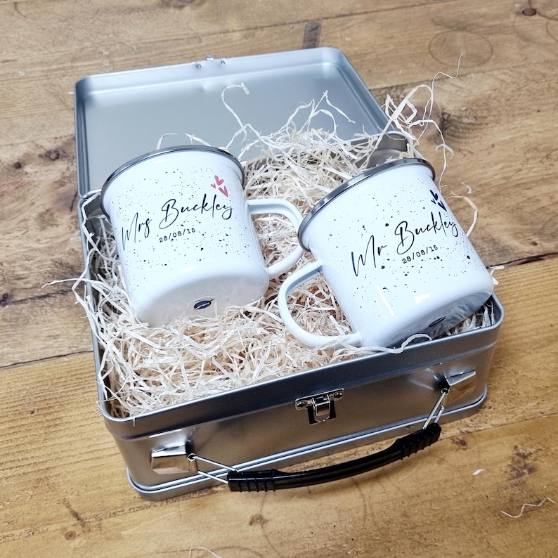 May include: Two white enamel mugs with black speckles and the names "Mrs Buckley" and "Mr Buckley" in script, along with the date "28/08/15". The mugs are inside a silver metal lunchbox filled with straw.