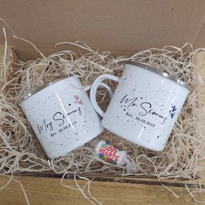 May include: Two white speckled enamel mugs with silver rims, personalized with "Mr Simms" and "Mrs Simms" and the date "Est. 22.02.2013". The mugs are in a cardboard box with straw, alongside a "Love Hearts" candy.