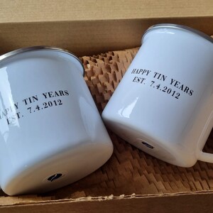Pode incluir: Duas canecas de esmalte branco com bordas prateadas. As canecas t&ecirc;m o texto "Happy Tin Years Est. 7.4.2012" impresso nelas.