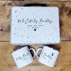 May include: A white metal lunchbox and two matching mugs with black speckles. The lunchbox has a black handle and the text "Mr & Mrs Buckley", "Happy "Tin" Years", and the date 28/08/15. The mugs have the names "Mr Buckley" and "Mrs Buckley" with the date.