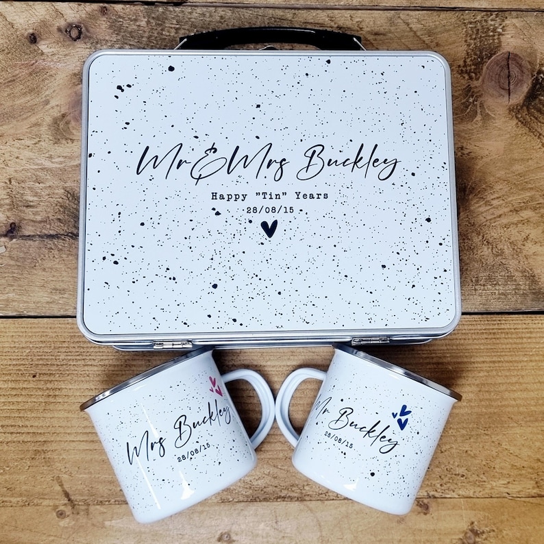 May include: A white metal lunchbox and two matching mugs with black speckles. The lunchbox has a black handle and the text "Mr & Mrs Buckley", "Happy "Tin" Years", and the date 28/08/15. The mugs have the names "Mr Buckley" and "Mrs Buckley" with the date.