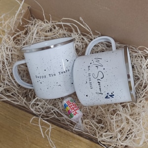 May include: Two white enamel mugs with black speckles and silver rims, nestled in a box with straw. One mug reads "Happy Tin Years," the other "Mr. Simms Est. 22.02.2013." A small candy roll with "Love Hearts" text is also in the box.